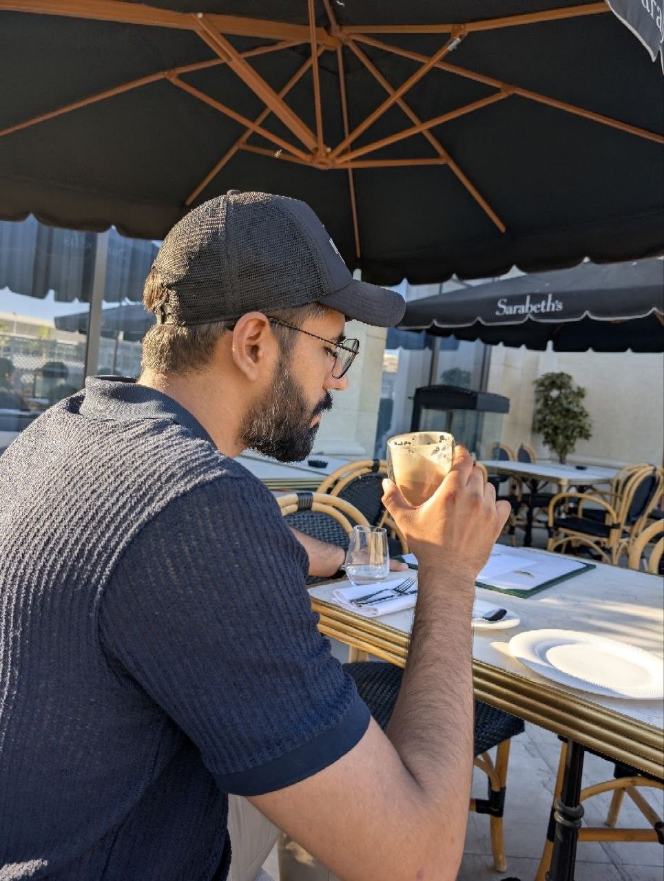 Faaiz on a café terrace, cap on, drink in hand